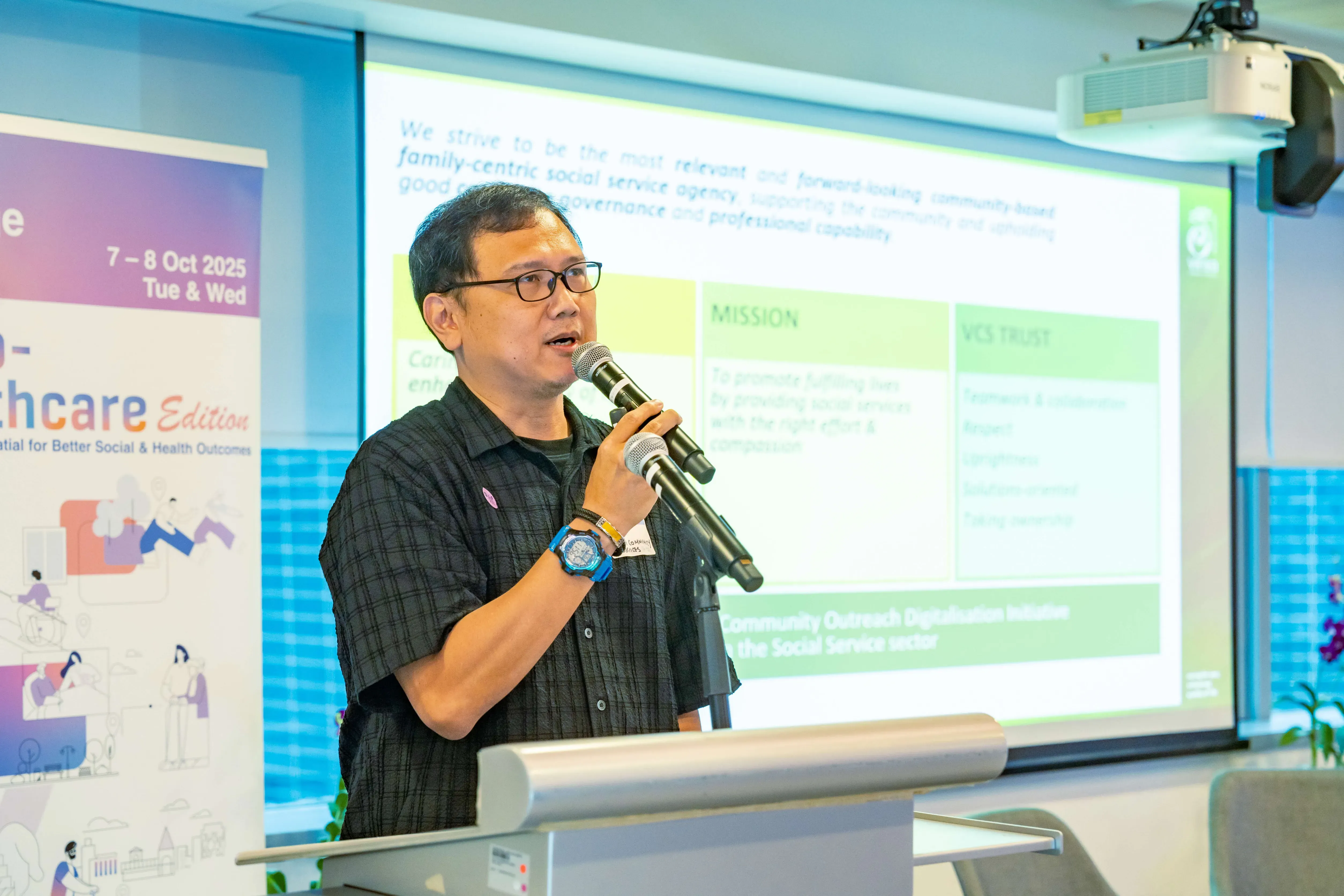 Man with glasses speaks into a microphone at a podium before a projected slide showing a mission statement.